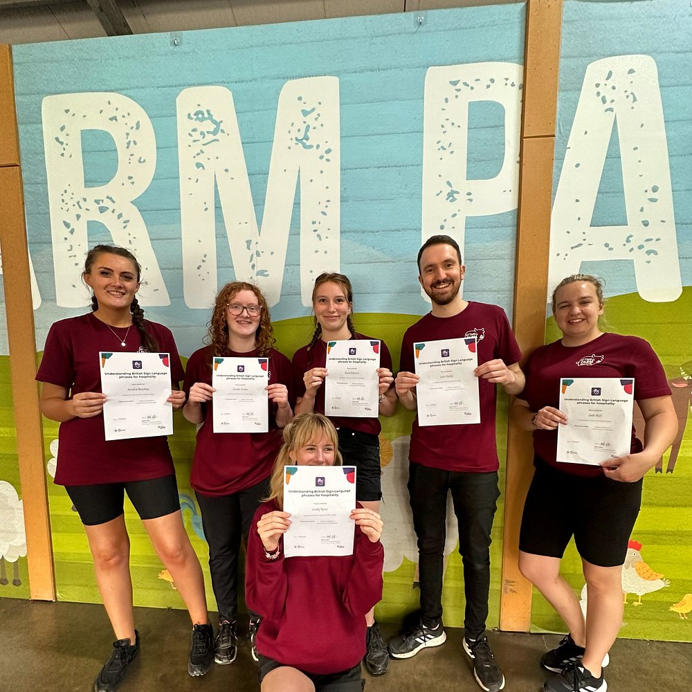 Six farm park staff members, dressed in matching maroon uniforms, proudly hold up their certificates after completing accessibility training. They are standing in front of a colorful wall mural with large text reading “FARM PARK,” smiling to celebrate their achievement.