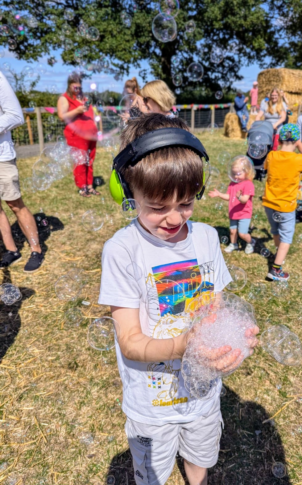 A young boy wearing bright green ear defenders smiles while playing with bubbles at an outdoor farm event. He is holding a handful of foam and surrounded by floating bubbles in the sunshine. Other children and adults are in the background, with hay bales, bunting, and trees visible, creating a lively and inclusive festival atmosphere.