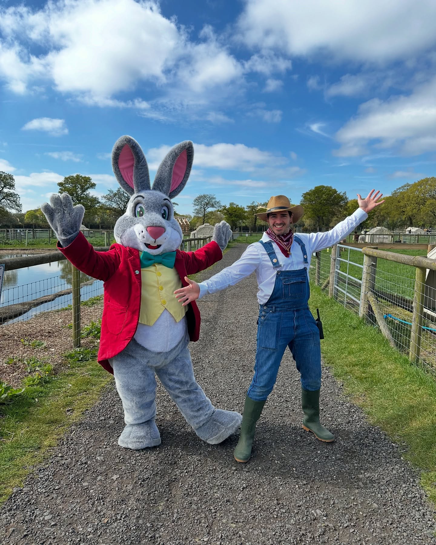 The Easter Bunny, wearing a red jacket, yellow waistcoat, and teal bow tie, stands smiling with arms raised beside a man dressed as a friendly farmer in denim overalls, green wellies, a straw hat, and red neckerchief. They are posing cheerfully on a farm path with fencing, grassy fields, and a pond in the background under a bright blue sky with scattered clouds.
