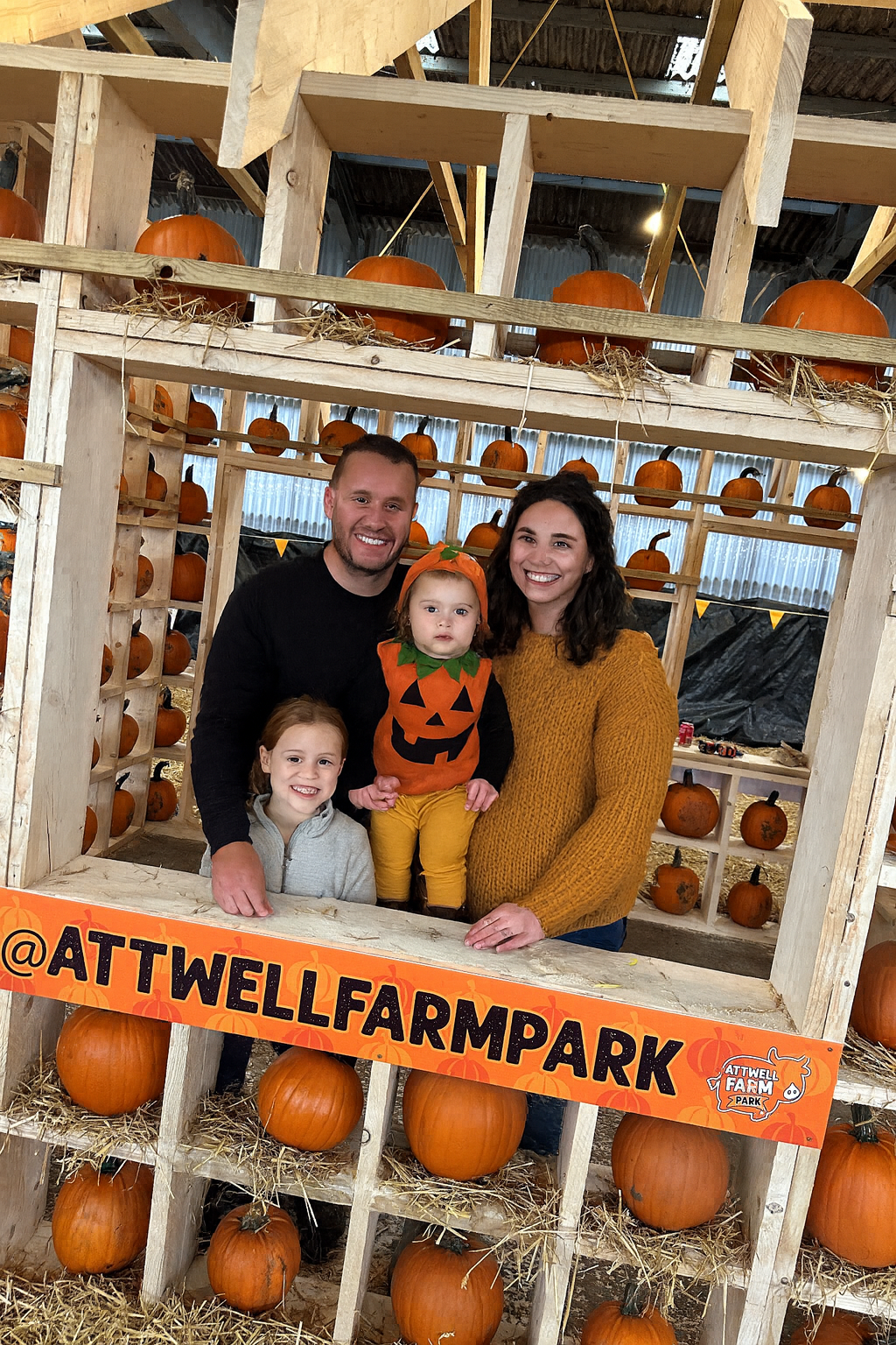 A family of four poses for a Halloween photo at Attwell Farm Park. They stand in a rustic wooden pumpkin display filled with orange pumpkins on shelves and straw. The father wears a black top, the mother a mustard jumper, and their two children smile in front, one dressed in a pumpkin-themed orange shirt and the other in a black outfit with a witch hat.