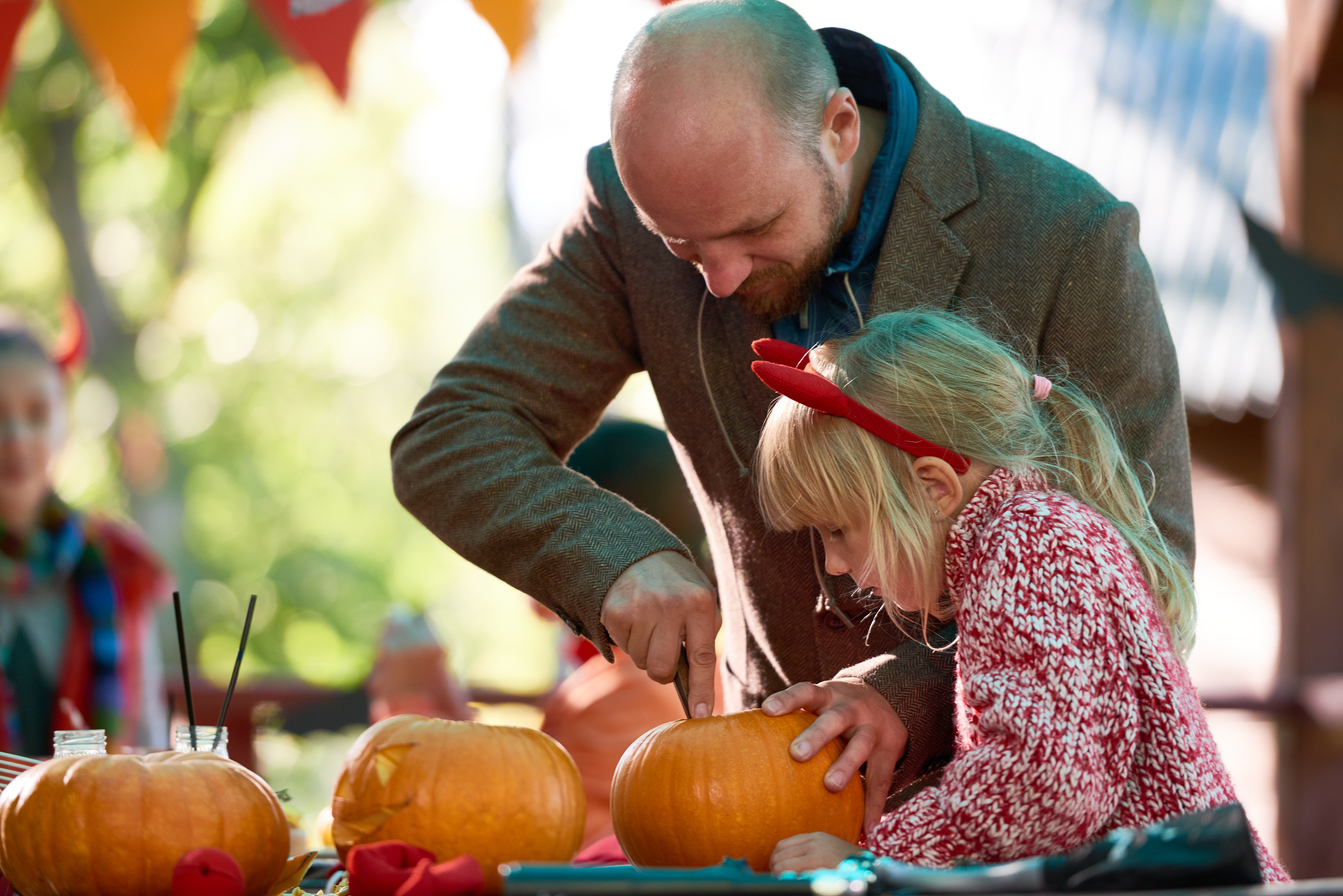 A man and a young girl carve pumpkins together at an outdoor carving table. The man carefully cuts into the top of a pumpkin while the girl, wearing a red headband and patterned jumper, steadies it with her hands. More pumpkins and carving tools sit on the table, and festive bunting hangs in the background, creating a warm autumn atmosphere