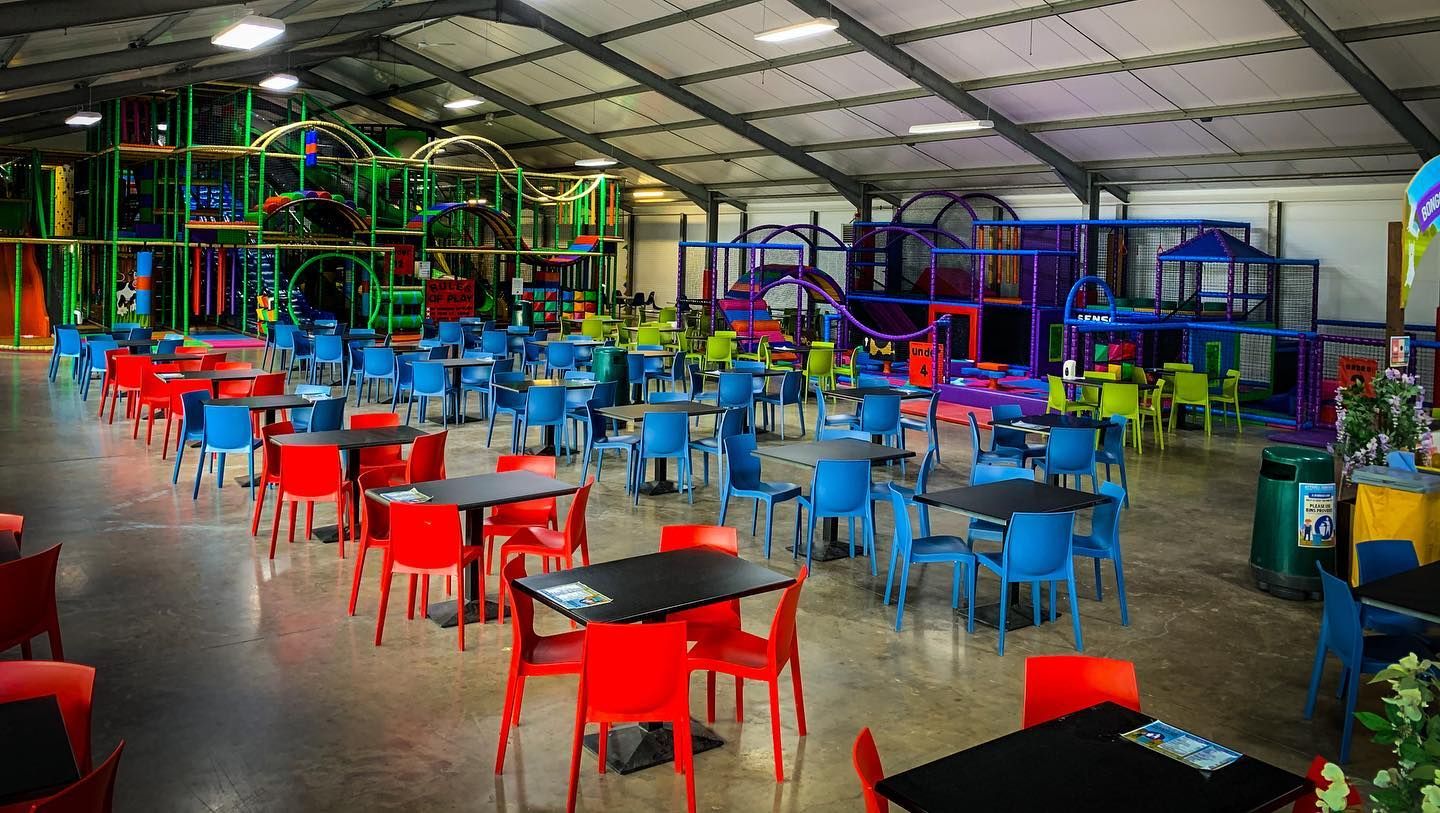 large indoor soft play barn with colourful play frames and seating. The play area features climbing structures, tunnels, and slides in bright green, purple, red, and blue. In the foreground, rows of square black tables are surrounded by red, blue, and green plastic chairs, arranged to provide plenty of seating for groups. The space is brightly lit with overhead lights, with wide walkways for easy access.