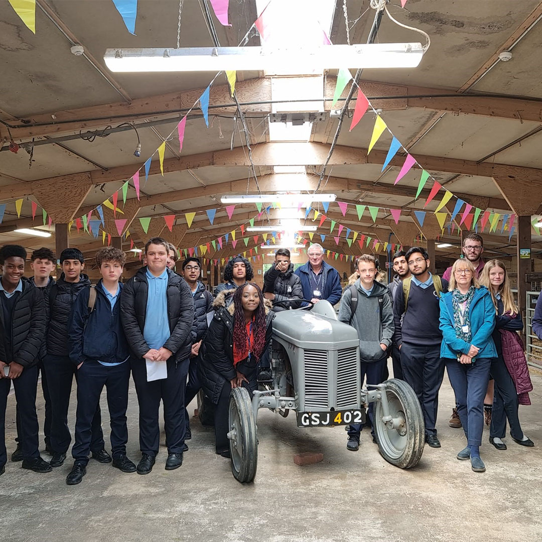 A large group of secondary school students and two adults pose together in a decorated barn. They are standing around a vintage grey tractor with the registration plate "GSJ 402." The barn ceiling is lined with colourful triangular bunting in yellow, pink, green, and blue. Most of the students are wearing school uniforms with dark trousers, shirts, and jackets, while the adults stand to the right and behind the tractor. Everyone is facing the camera, smiling or looking relaxed.