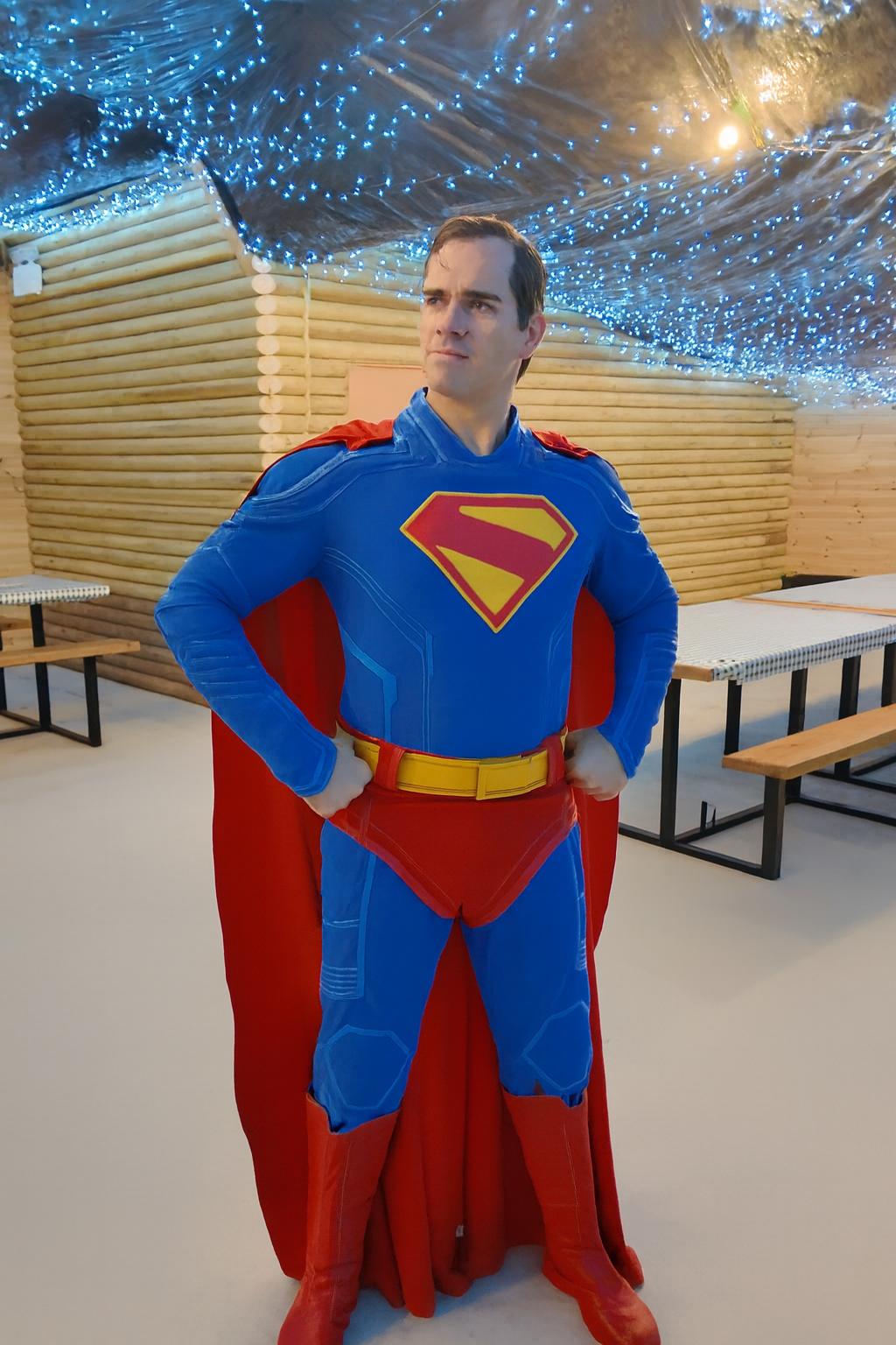 A man dressed as a superhero in a blue suit with a red cape, red boots, and a yellow belt stands confidently in a wooden party room with picnic-style tables. The ceiling is decorated with strings of blue fairy lights, creating a starry-night effect. The floor is smooth and step-free, making the space wheelchair accessible.