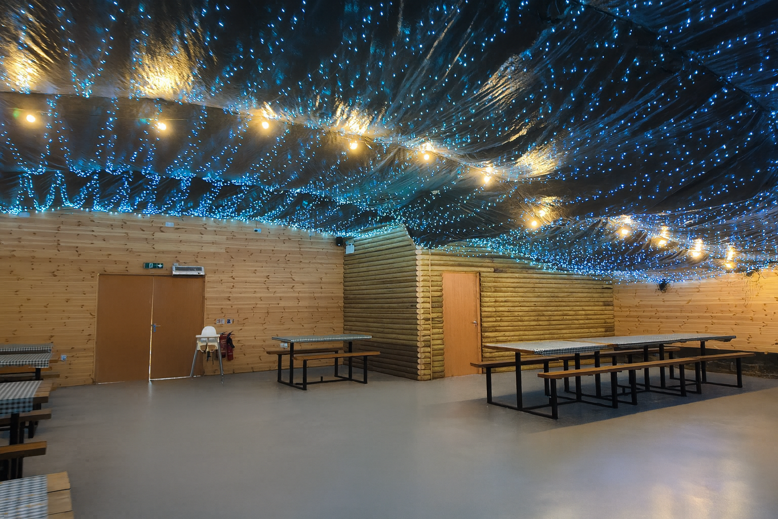 A spacious wooden party room with log-cabin style walls and long picnic-style tables with benches. The ceiling is draped in dark fabric, decorated with glowing blue fairy lights and warm string bulbs, creating a magical, starry-night effect. The floor is smooth and step-free for easy wheelchair access. A high chair and fire extinguisher are visible near the door, showing family-friendly and safety considerations.