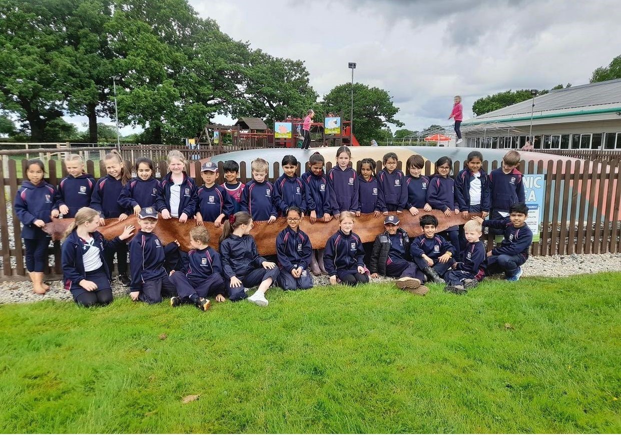 A large group of school children in matching navy tracksuits pose outdoors in front of a wooden fence, each holding onto a long, brown snake skin stretched out in front of them. Some children are standing while others are kneeling or sitting on the grass. Behind the fence is a play area with colourful signs, trees, and a giant inflatable jumping pillow where other children are playing. The group looks engaged and cheerful, showing the scale of the snake skin.