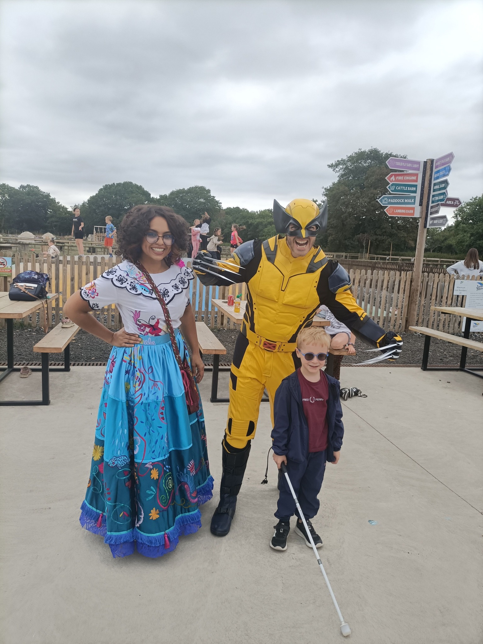 A young boy with vision impairment, holding a white cane and wearing sunglasses, smiles while standing with two costumed characters at a farm park. On his left is a woman in a colorful embroidered dress with floral patterns, and on his right is a man in a bright yellow superhero costume with black details and a mask. They are outdoors on a paved area with picnic benches, fencing, and directional signs visible in the background.
