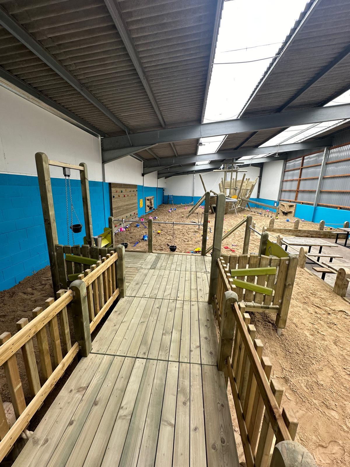 An indoor sandpit play area with a wide wooden ramp and handrails providing wheelchair access. The ramp leads into a spacious play zone with sand, climbing structures, and swings, designed for inclusive play. Bright blue walls and natural light from skylights make the area welcoming and accessible for children of all abilities.